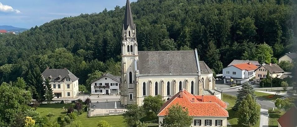 Ein Luftbild einer Kirche, umgeben von Häusern und Grünflächen, vor einer Kulisse von Hügeln und Bäumen.
