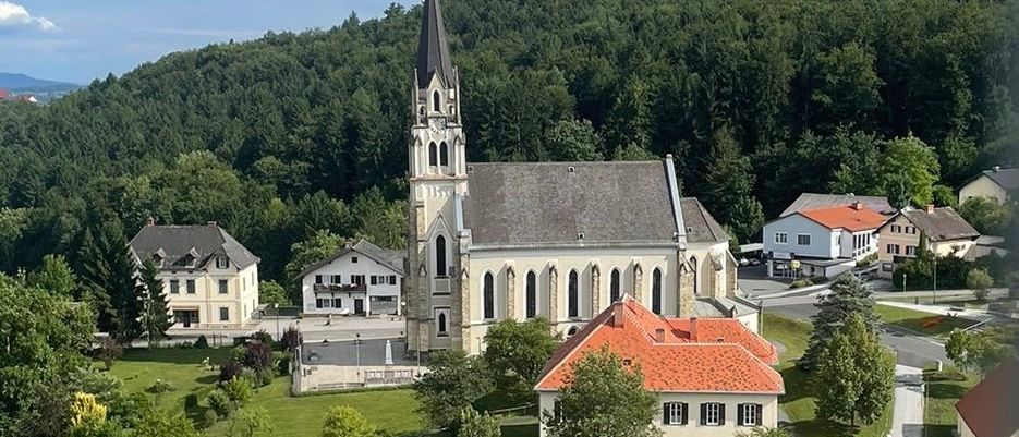 Ein Luftbild einer Kirche, umgeben von Bäumen und Häusern, mit einem rotgedeckten Gebäude im Vordergrund.