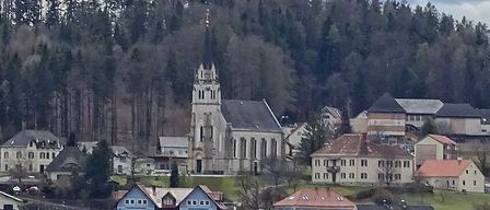 Eine kleine Stadt mit vielen Häusern und einer Kirche, umgeben von dichtem Wald. Die Kirche hat einen hohen Turm und ein graues Dach.