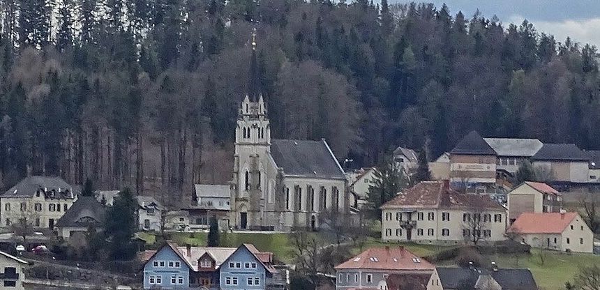 Ein Dorf mit verschiedenen Häusern, einer großen Kirche mit einem Turm, umgeben von Grün und dichtem Wald.