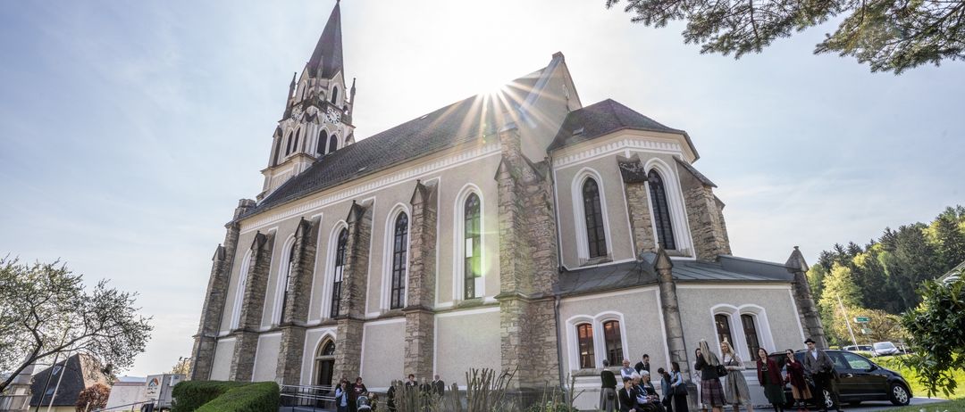 Eine große gotische Kirche mit Glockenturm und Spitzdach. Mehrere Menschen sind draußen versammelt, einige stehen und andere sitzen auf Stufen. Die Sonne scheint hell.