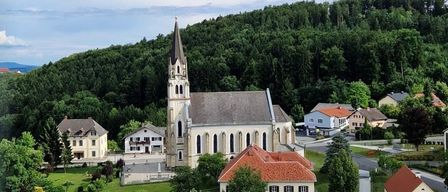 Luftaufnahme einer Kirche mit einem Turm, umgeben von Häusern, Bäumen und einem Berg unter einem bewölkten Himmel.