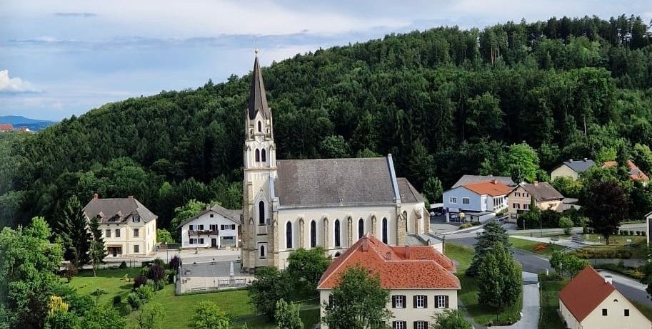Luftaufnahme einer Kirche umgeben von Häusern und Grünflächen, eingebettet in einem Tal mit blauem Himmel und Wolken.