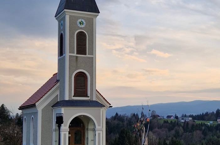 Eine kleine Kapelle mit einem spitzen Turm, vor dem Hintergrund von Hügeln und einem Sonnenuntergangshimmel. Die Kapelle hat ein rotes Dach, eine Uhr und eine Holztür. Es gibt einen Zaun um die Kapelle, und eine Statue in der Nähe.