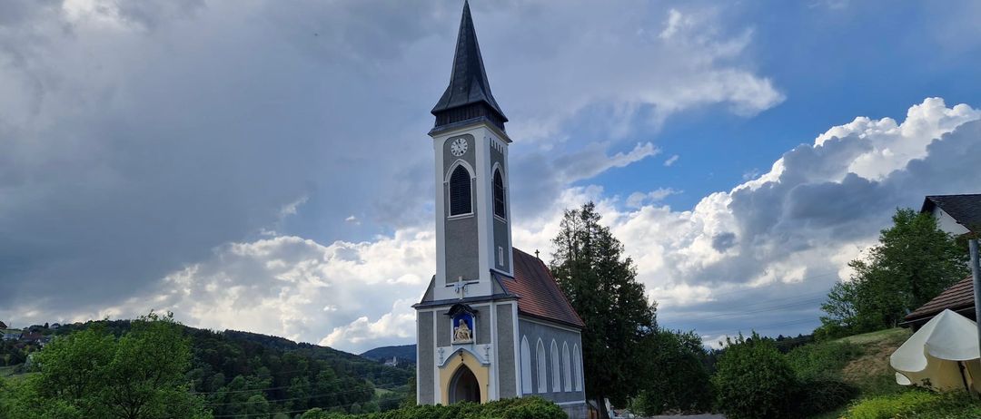 Eine Kirche mit einem Turm und einer Uhr steht unter einem bewölkten Himmel. Das Gebäude hat ein rotes Dach und ein Kreuz an der Spitze.