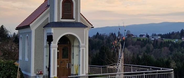 A small chapel with a steeple and a clock stands on a hill, surrounded by a metal fence and a small garden. The chapel is set against a backdrop of mountains and a cloudy sky.