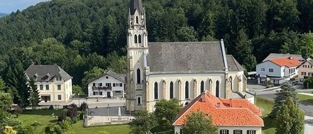 Luftaufnahme einer Kirche mit Turm und rotem Dach, umgeben von Häusern und Bäumen in einem bergigen Gebiet.