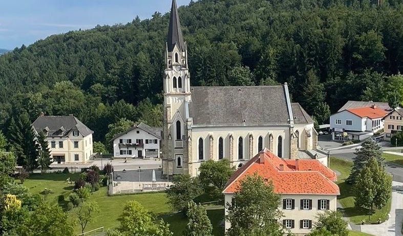 Luftaufnahme einer Kirche mit Turm und rotem Dach, umgeben von Häusern und Bäumen in einem bergigen Gebiet.