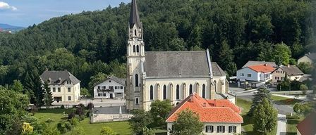 Luftaufnahme einer Kirche mit einem Turm, umgeben von Häusern und Bäumen, mit einem Berg im Hintergrund.