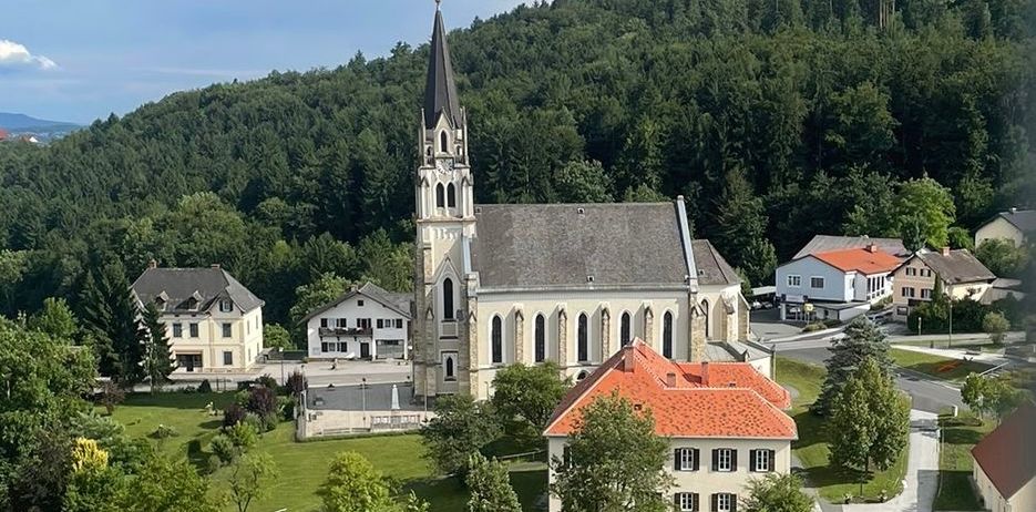 Luftaufnahme einer Kirche mit einem Turm, umgeben von Häusern und Bäumen, mit einem Berg im Hintergrund.
