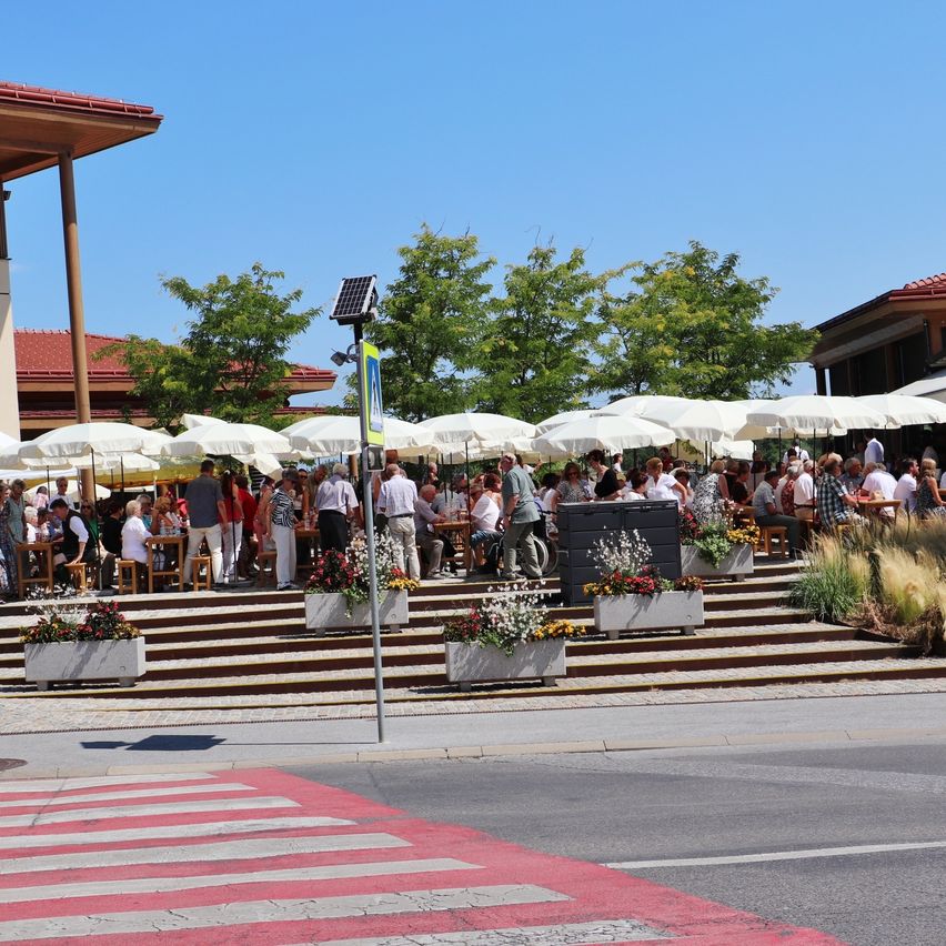 Eine Gruppe von Menschen trifft sich in einem Restaurant im Freien und genießt den sonnigen Tag. Das Restaurant ist mit weißen Sonnenschirmen geschmückt, und der Bereich ist von Bäumen umgeben.