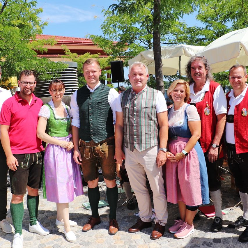 Eine Gruppe von Menschen in traditioneller deutscher Kleidung steht lächelnd für ein Foto im Freien. Hinter ihnen sind Bäume und ein Schirm.