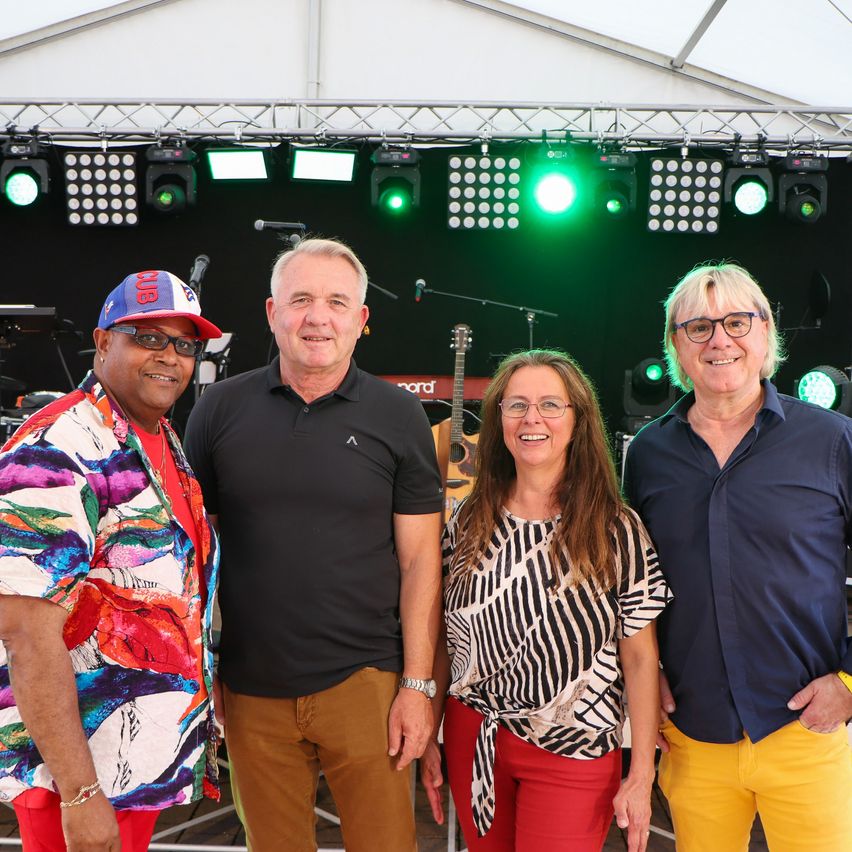 Vier Personen stehen lächelnd auf einer Bühne, mit grünen Bühnenlichtern im Hintergrund und Musikinstrumenten in der Nähe.