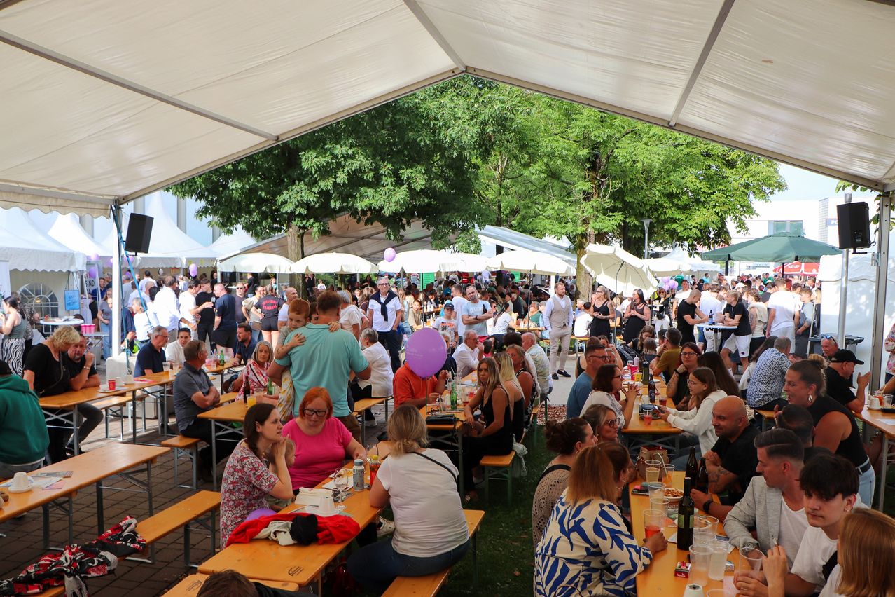 Eine große Gruppe von Menschen unter einem Zelt, die Essen und Getränke genießen. Einige sitzen, andere stehen und gehen. Die Umgebung ist im Freien mit Bäumen im Hintergrund.