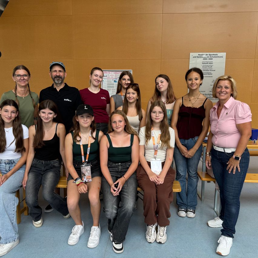 Eine Gruppe von Personen, darunter Erwachsene und junge Frauen, stehen und sitzen auf Bänken in einem Raum. Sie scheinen für ein Foto zu posieren. Die Frau auf der rechten Seite lächelt und hat die Hände in die Taille gestemmt.