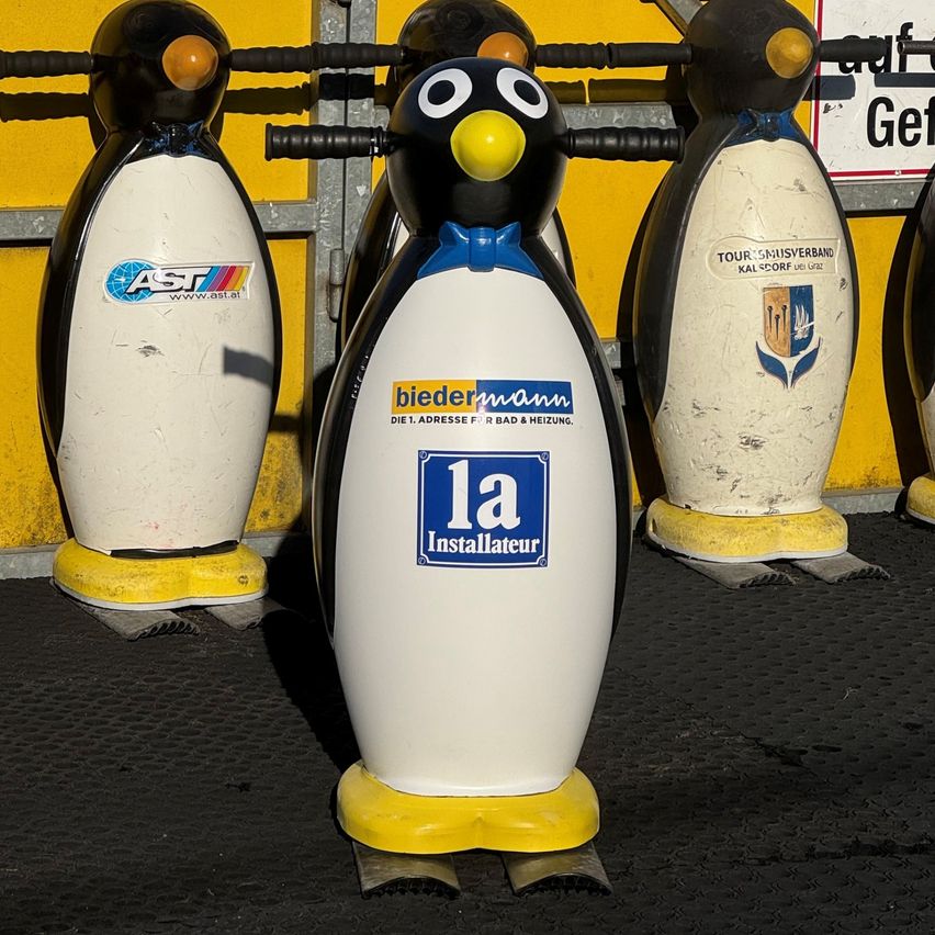 Vier Pinguinskulpturen sind auf einer schwarzen Oberfläche aufgereiht. Sie haben gelbe Füße und schwarze Griffe. Eine hat einen blauen Aufkleber mit der Aufschrift 'la Installateur'.