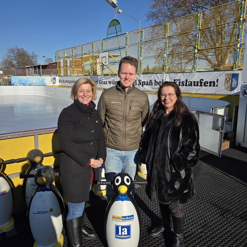 Drei Personen stehen neben Pinguin-Skulpturen auf einer Eisbahn und lächeln für ein Foto. Hinter ihnen befindet sich ein Banner mit deutschem Text, und die Eisbahn ist mit gelben Barrieren eingezäunt.