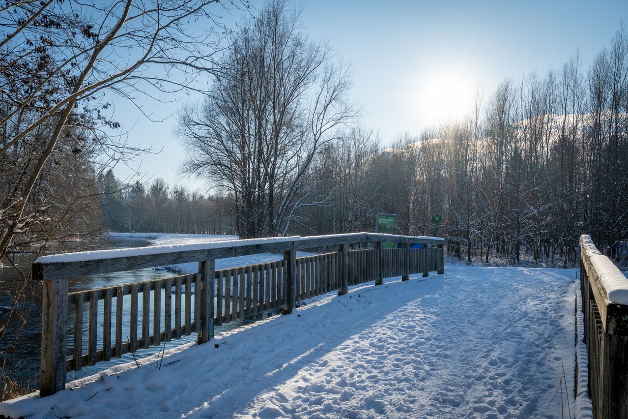 Ein verschneiter Holzzaun steht neben einem zugefrorenen Fluss, mit kahlen Bäumen und einer strahlenden Sonne im Hintergrund.