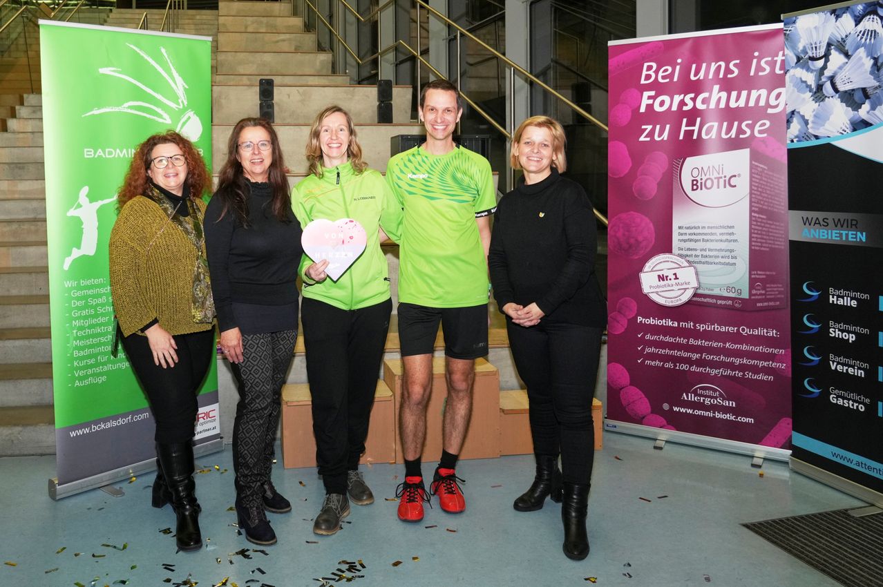 Fünf Personen stehen auf einer Bühne und lächeln für ein Foto. Ein Mann in der Mitte hält ein herzförmiges Objekt. Hinter ihnen befinden sich zwei Banner mit Text. Die Treppe und der Boden sind mit Konfetti bedeckt.