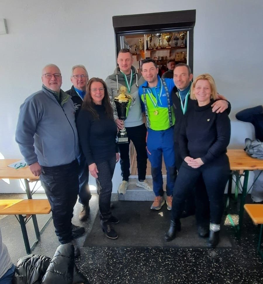 Eine Gruppe von Menschen steht zusammen, lächelt und posiert für ein Foto. Sie tragen Winterkleidung und Medaillen. Die Person in der Mitte hält eine Trophäe. Dahinter steht ein Tisch mit Bänken.