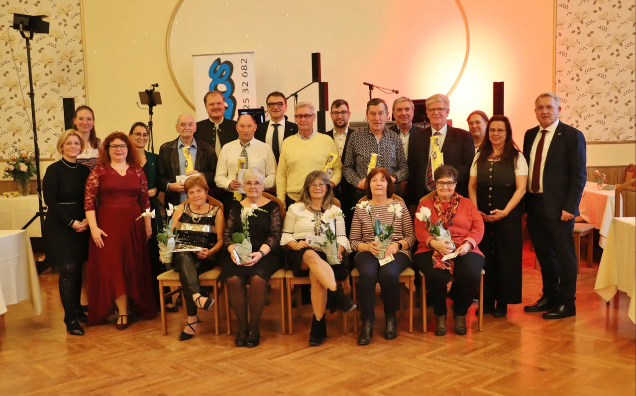 Eine Gruppe von Menschen, sowohl Männer als auch Frauen, ist zu einer formellen Veranstaltung, möglicherweise einer Preisverleihung, versammelt. Sie sitzen auf Stühlen, halten Blumen und scheinen für ein Foto zu posieren. Einige Personen tragen Krawatten und Brillen.