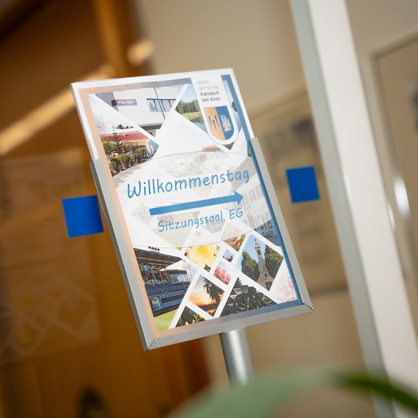 Ein Prospekt mit dem Titel 'Willkommenstag' wird auf einem Metallständer mit blauen Tabs ausgestellt. Das Bild zeigt verschiedene Fotos von Gebäuden, einem Gebäude mit Polizeischild und einem Sonnenuntergang.