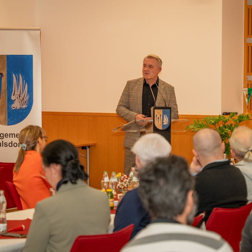 Ein Mann spricht an einem Podium in einem Konferenzraum mit einem Mikrofon. Er steht vor einem sitzenden Publikum mit einem Tisch mit Getränken und Essen. Ein Schild und eine Pflanze sind hinter ihm.