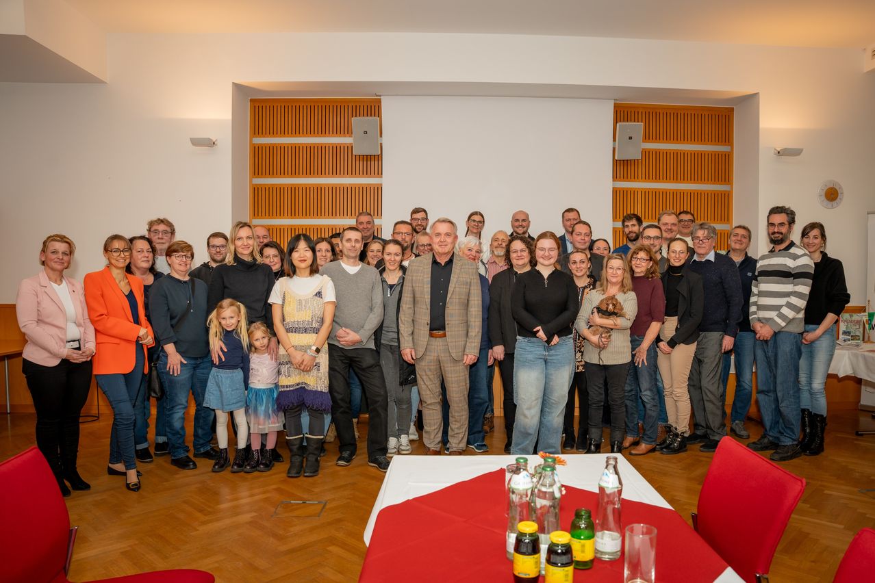 Eine Gruppe von Menschen steht in einem Raum für ein Foto. Ein Mann in einem Anzug steht vor einem roten Tisch mit Flaschen. Hinter ihm hält eine Frau einen Hund. Holzpaneele sind an der Wand.