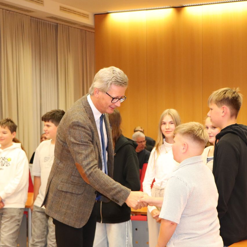 Ein Mann in formeller Kleidung schüttelt einem jungen Jungen in einem hellen Hemd die Hand. Mehrere andere Kinder und Erwachsene sind anwesend und beobachten. Der Raum ist gut beleuchtet.