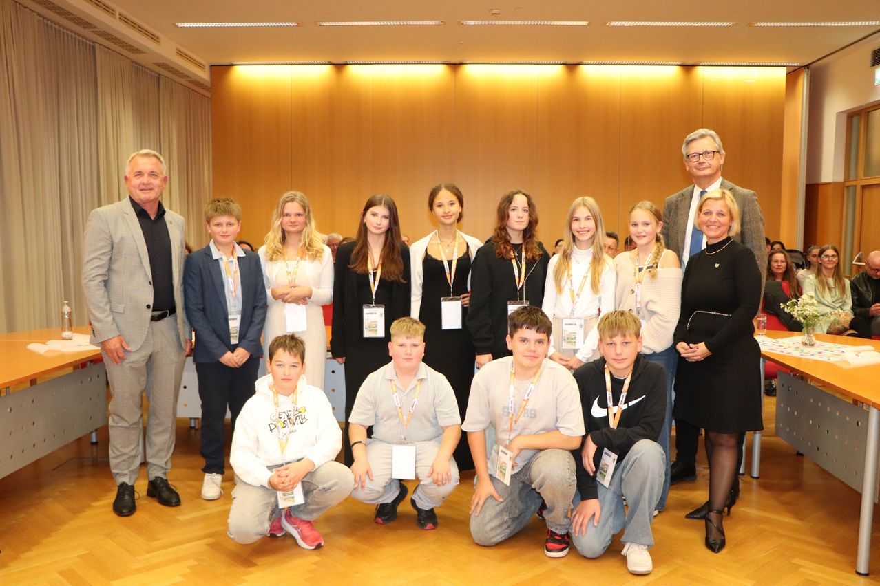 Eine Gruppe junger Menschen und Erwachsene stehen und knien in einem Raum mit Holzböden und Wänden. Sie tragen Namensschilder und scheinen für ein Foto zu posieren.