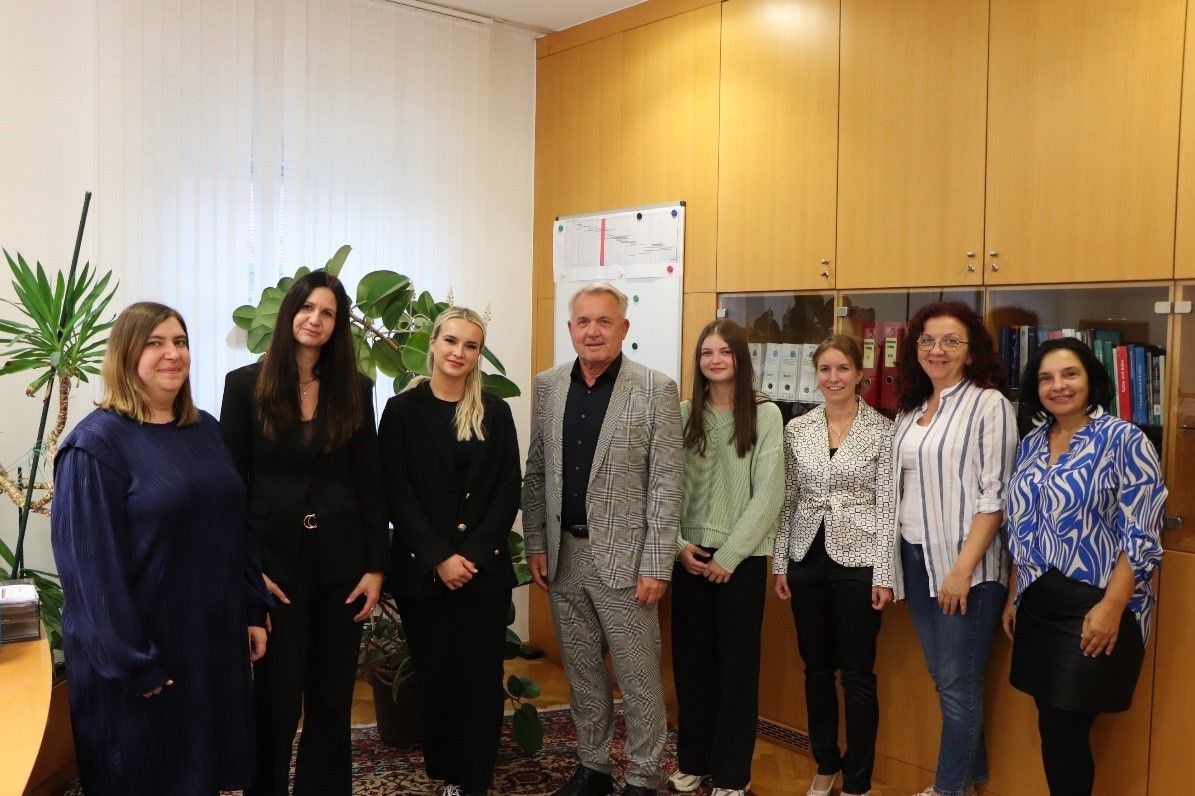 Sieben Erwachsene, darunter drei Frauen und ein Mann im Anzug, posieren für ein Foto in einem Büro. Dahinter steht eine Pflanze und eine Tafel an der Wand.