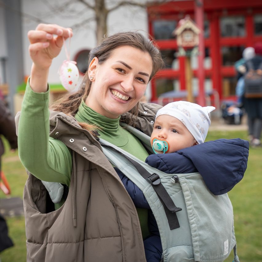 A woman in a green coat holds an Easter egg and smiles, carrying a baby in a blue carrier with a pacifier. They stand on a grassy area.