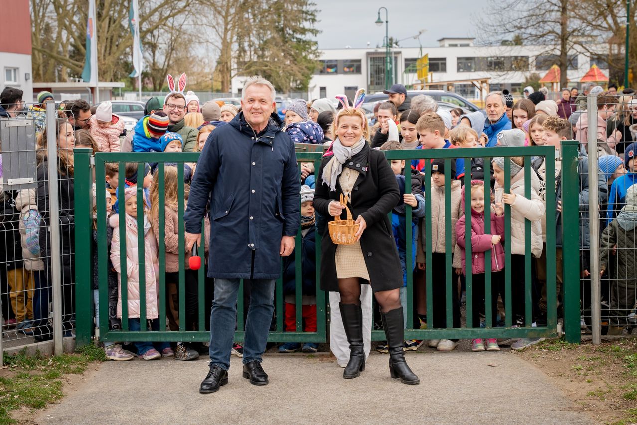 A group of adults and children stand behind a green fence. A man and a woman are holding a basket with Easter eggs.