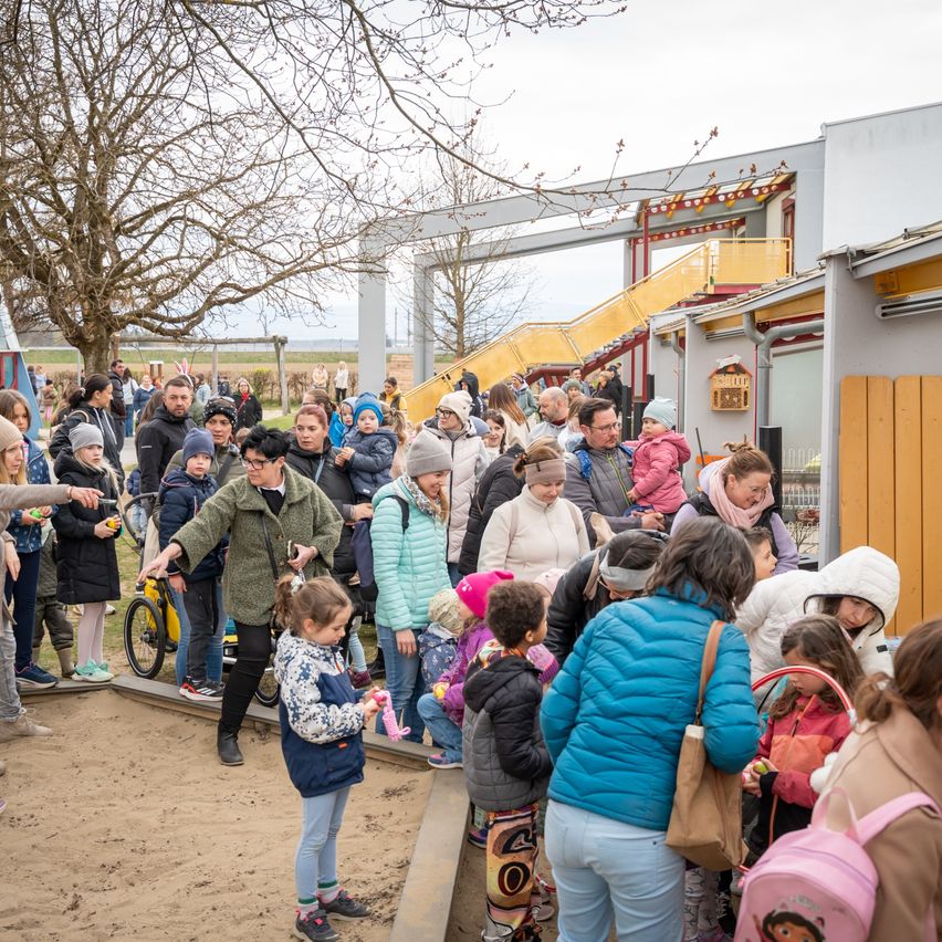 A crowd of adults and children gather outdoors, some in line, others chatting, near a play area with slides. The area is surrounded by trees, and some individuals hold Easter eggs.