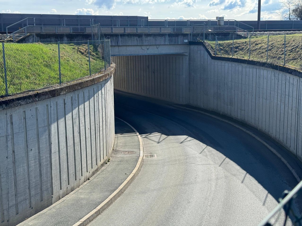A tunnel entrance with concrete walls and a grassy hill above it. The road is empty, and shadows are cast on the road.