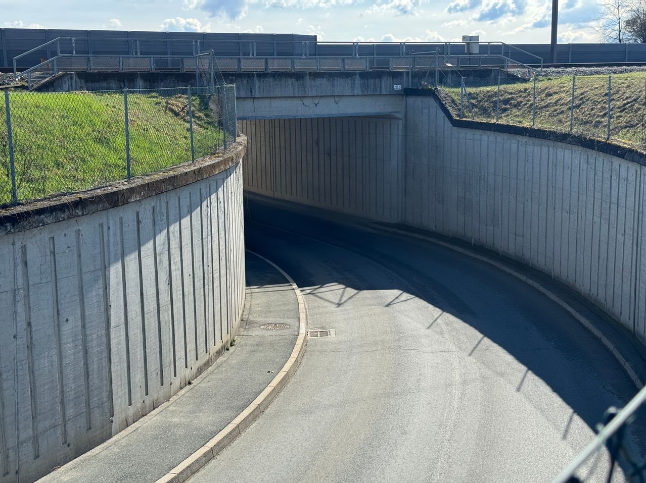 A road tunnel with a concrete arch, leading into a large, open area. Grassy hills are on either side of the tunnel, with a bridge above.