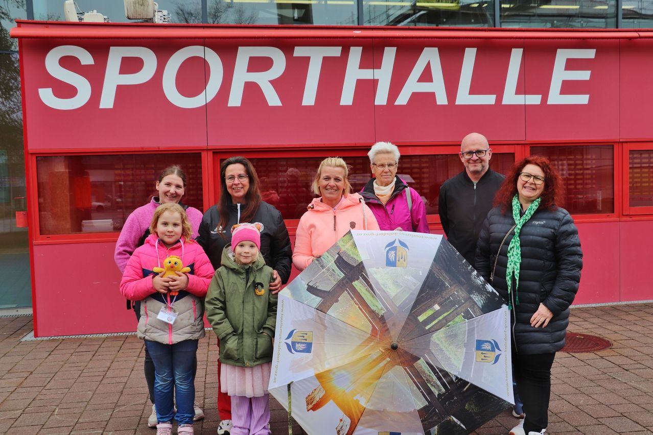 A group of adults and children stands in front of a red building labeled 'PORTHALL', holding an umbrella with various designs and a girl with a stuffed toy.