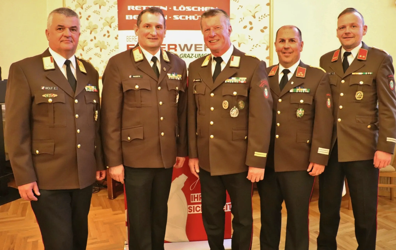 Four men in military uniforms stand together in a formal setting. They are smiling and looking at the camera. Behind them is a banner with text.
