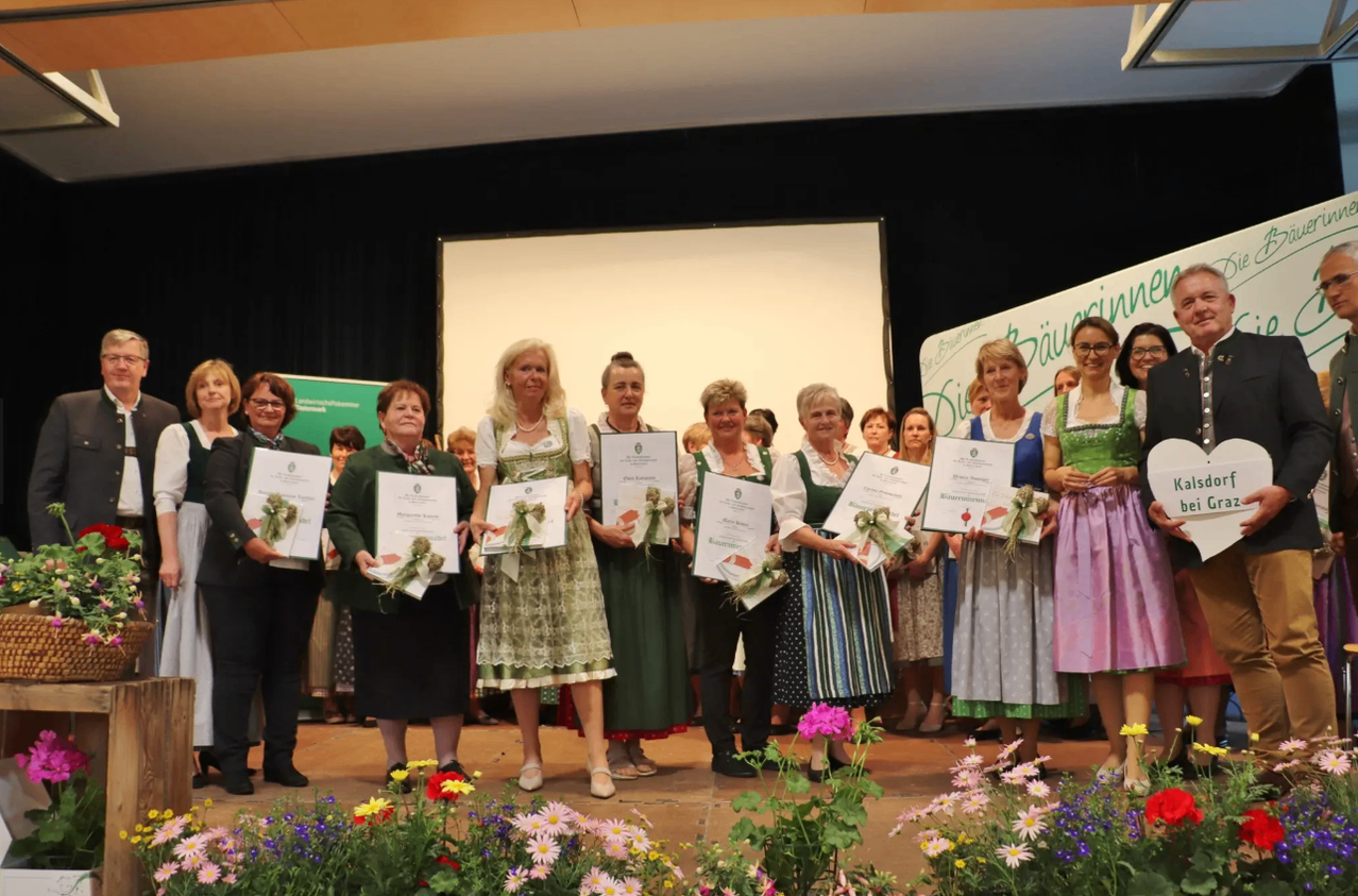 A group of women in traditional Bavarian clothing holding certificates and bouquets stand on stage, surrounded by flowers.
