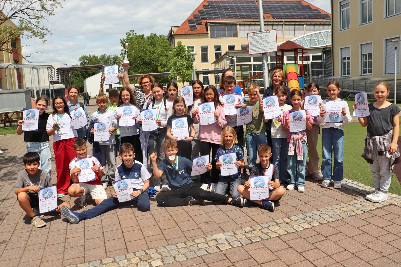 Eine Gruppe von Kindern und Erwachsenen, die Urkunden halten, posiert für ein Foto mit einer Trophäe vor einem Gebäude mit Solarpanelen.