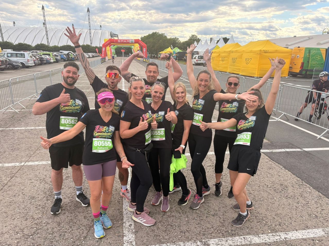 Gruppe von Läufern, die schwarze T-Shirts und nummerierte Startnummern tragen. Sie stehen in einer Reihe mit erhobenen Armen und lächeln für die Kamera. Sie befinden sich an der Startlinie eines Marathons.