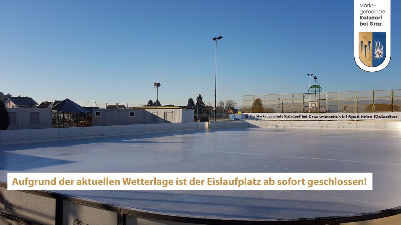 An empty ice rink with a clear sky and street lights. A building with a fence is nearby. The ice rink has a banner with text.