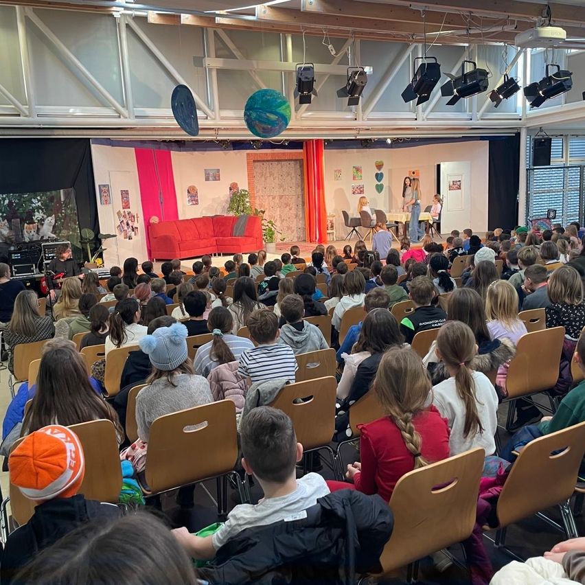 Ein großer Raum mit Stuhlreihen, die auf eine Bühne gerichtet sind, auf der zwei Frauen vor einem roten Sofa auftreten. Scheinwerfer hängen von der Decke ab.