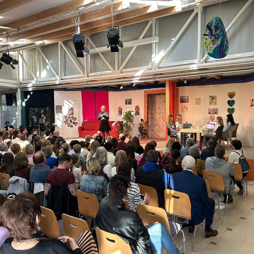 Ein Raum mit einer Bühne, auf der eine Frau steht und spricht, mit einem Tisch und Stühlen aufgestellt. Unten sitzen Menschen in Stühlen, dem Bühnenbereich zugewandt, mit einem roten Sofa und Pflanzen im Hintergrund.