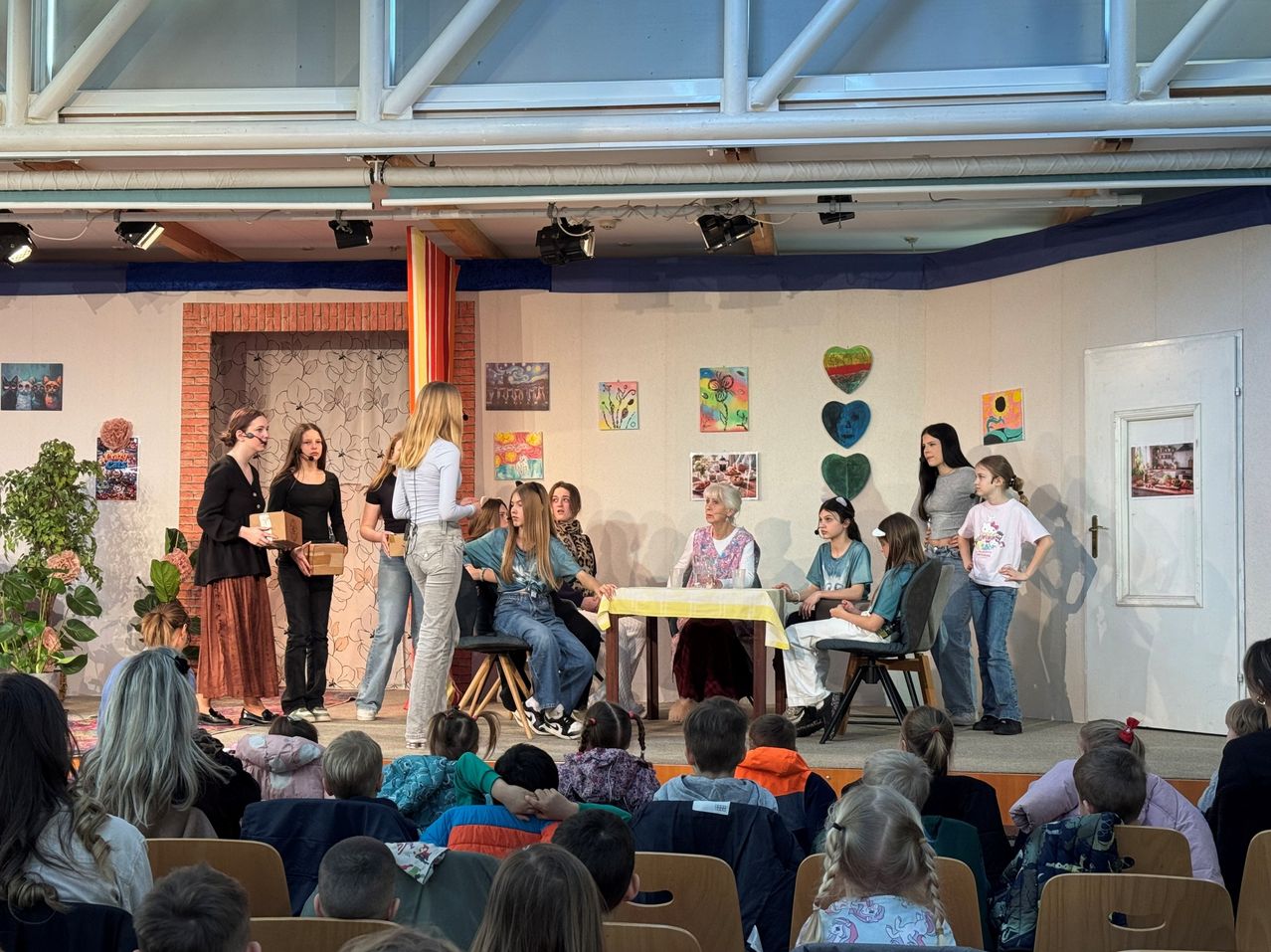 Eine Gruppe von Menschen auf der Bühne führt ein Theaterstück auf, das von einem Publikum unten beobachtet wird. Die Bühne ist mit Gemälden geschmückt.