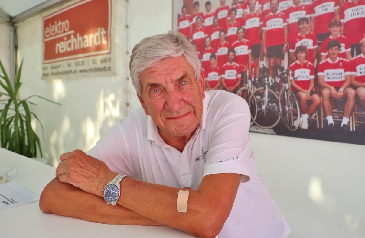 Ein älterer Mann mit weißen Haaren und einem Verband am rechten Arm sitzt vor einer Wand mit einem Poster von Radfahrern in roten Trikots.