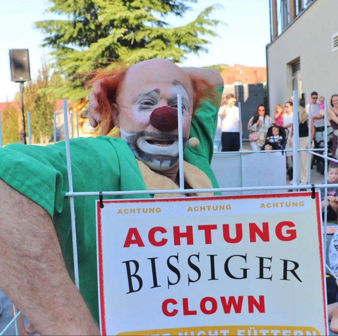 Eine Person, die als Clown verkleidet ist und ein Schild mit der Aufschrift 'ACHTUNG BISSIGER CLOWN' vor einer Menschenmenge hält.