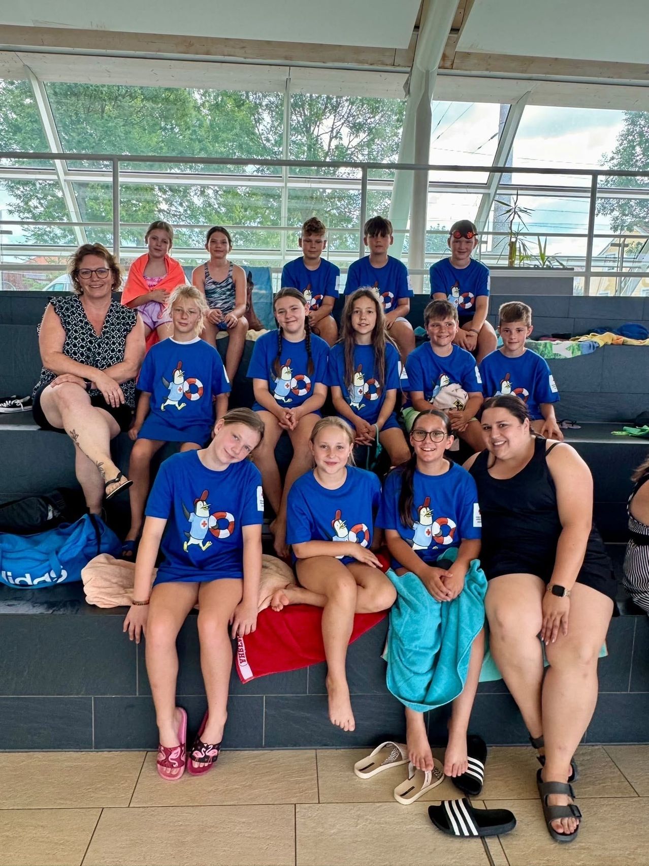 Eine Gruppe von Kindern und Erwachsenen in passenden blauen T-Shirts mit einem Pinguin-Design sitzen auf Bänken in einem Außenpoolbereich. Die Erwachsenen lächeln und die Kinder sehen glücklich aus.