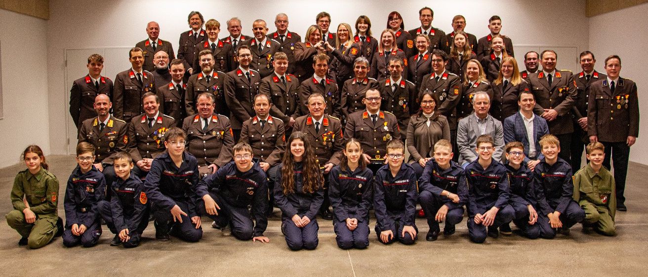 Eine Gruppenaufnahme von Personen in Militäruniformen, darunter einige in Kadettenkleidung, posieren für ein Foto in einem Raum mit weißen Wänden und Deckenleuchten.