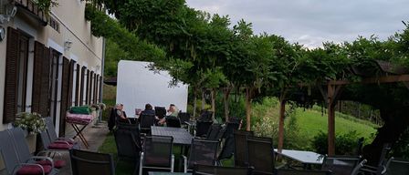 An outdoor dining area with tables and chairs surrounded by greenery, featuring a building with windows and awnings, under a cloudy sky.